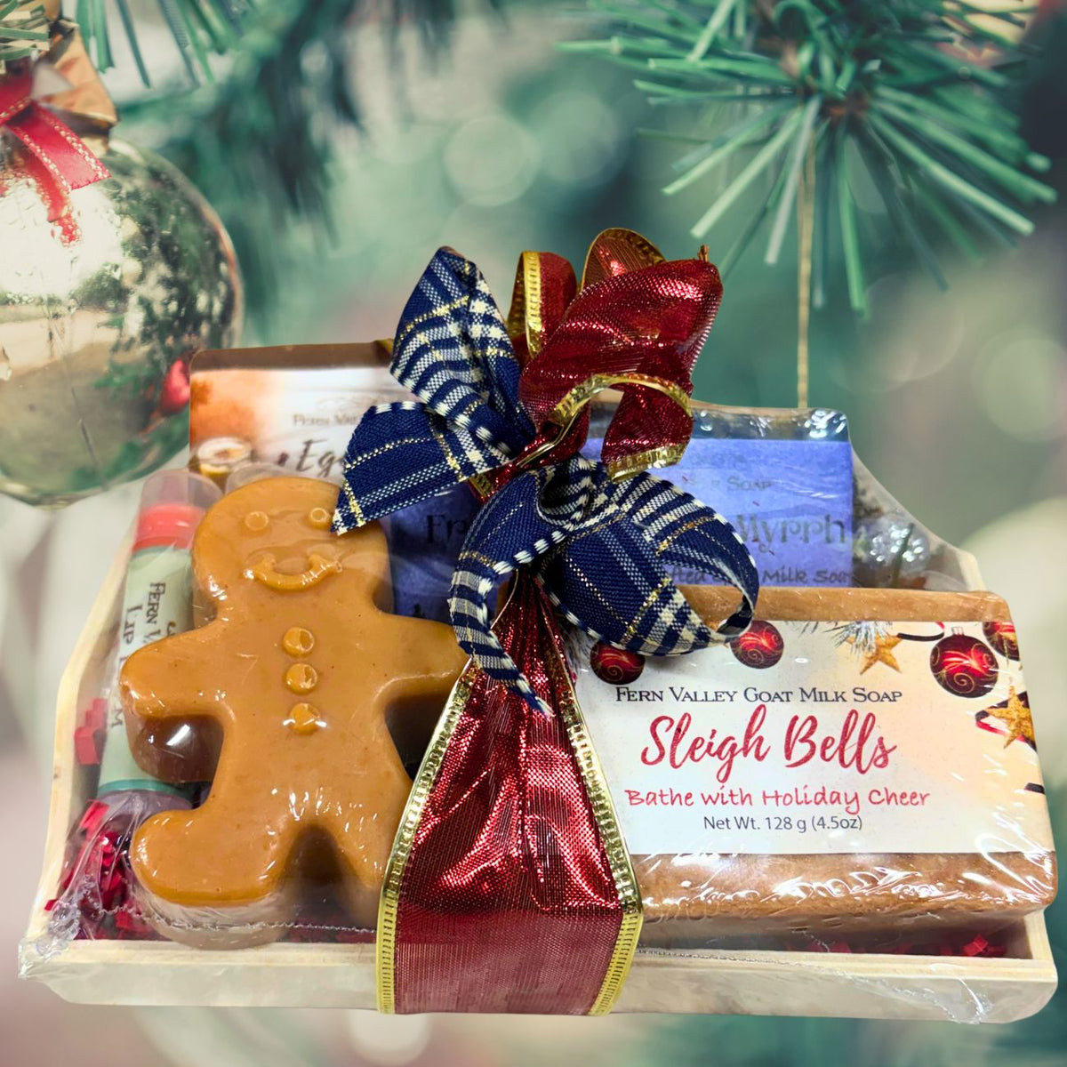 A festive holiday gift tray wrapped with a shimmering blue and red plaid bow, featuring a gingerbread man novelty soap and a bar of Sleigh Bells goat milk soap.