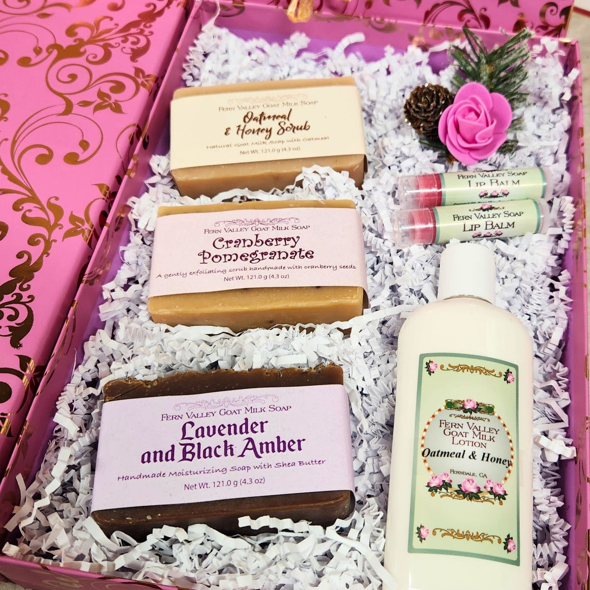 An open premium holiday gift box filled with clean white crinkle paper, three labeled Fern Valley goat milk soaps (Oatmeal & Honey, Lavender Black Amber, Cranberry Pomegranate), a bottle of lotion, and two lip balms.