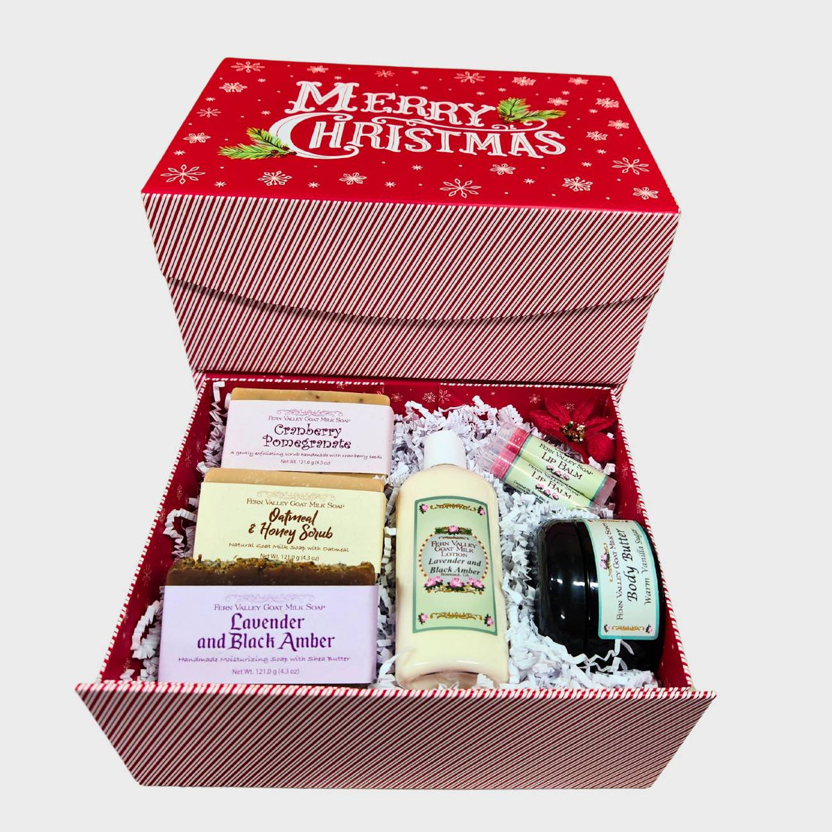 An open red and white striped Christmas gift box neatly filled with white crinkle paper, three Fern Valley goat milk soap bars (Cranberry Pomegranate, Oatmeal & Honey, Lavender and Black Amber), a bottle of lotion, a jar of body butter, and two lip balms.