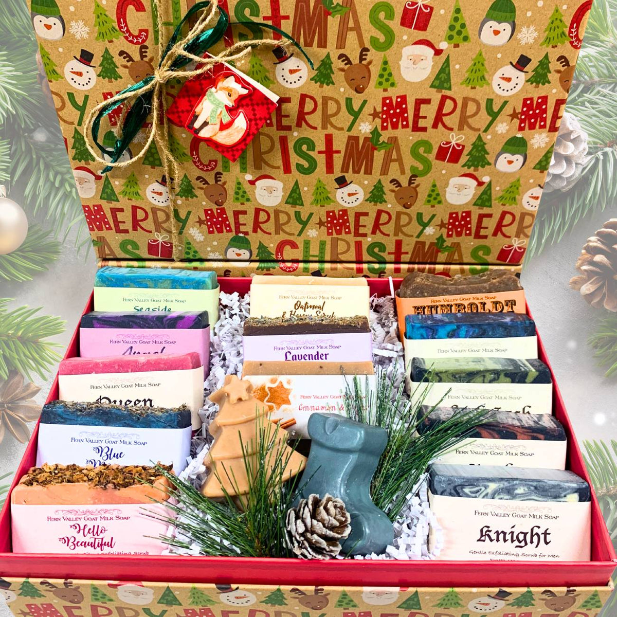 A Christmas-themed gift box filled with assorted soap bars, wrapped in a red and green patterned box with holiday illustrations. The box is decorated with a wooden reindeer ornament and surrounded by pine branches and a pinecone, with a green pine tree backdrop.
