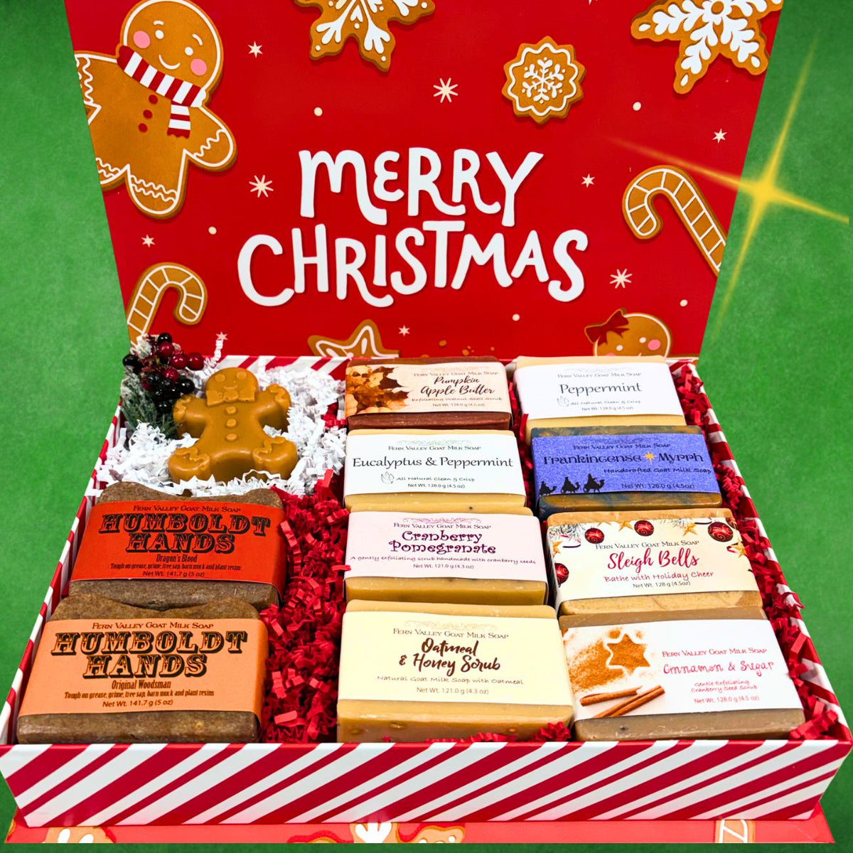 A Christmas-themed gift box filled with assorted artisanal soap bars. The open red-and-white striped box features a festive red lid decorated with gingerbread cookies, candy canes, and the words &