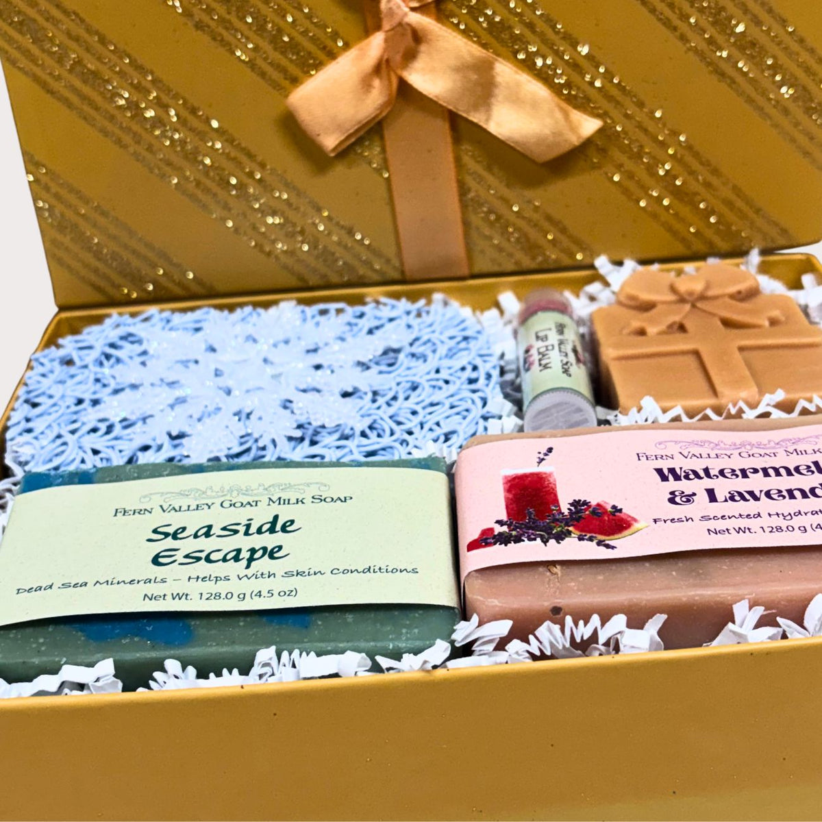 Close-up shot of an open gold gift box containing Seaside Escape and Watermelon & Lavender handmade goat milk soap bars, a clear tube of all-natural lip balm, and a molded holiday present soap shape, on a clean white background.
