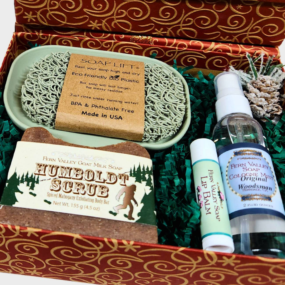 An open copper and gold patterned Christmas gift box filled with green crinkle paper, displaying a Humboldt Scrub goat milk soap bar, a 2 oz Original Woodsman cologne spray, a lip balm, and a sage green self-draining soap dish with a matching soap lift.