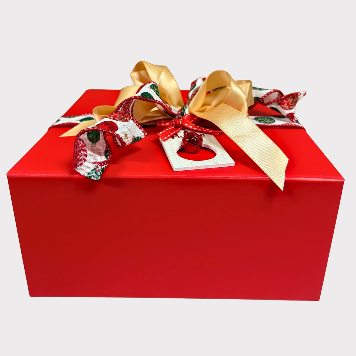 A closed square crimson red gift box elegantly tied with a gold satin bow, a red and green holly patterned ribbon, and a tiny red jingle bell ornament tag against a white background.