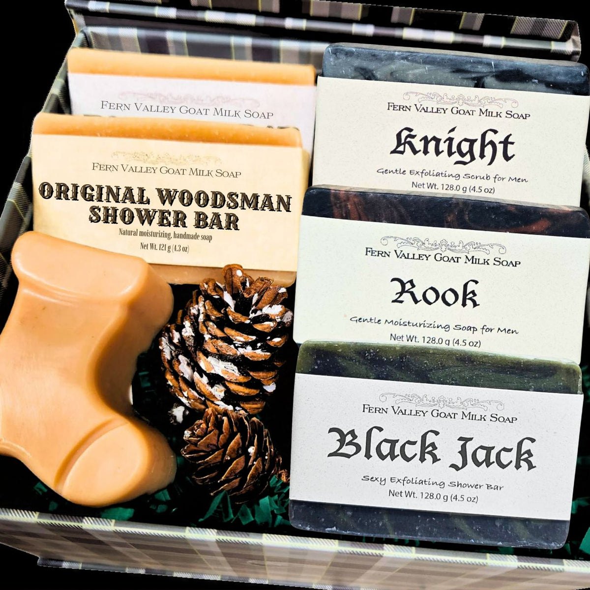 A plaid holiday gift box filled with Fern Valley Goat Milk Soaps for men, including Original Woodsman, Rook, and Black Jack bars, next to a boot-shaped soap and pinecones.