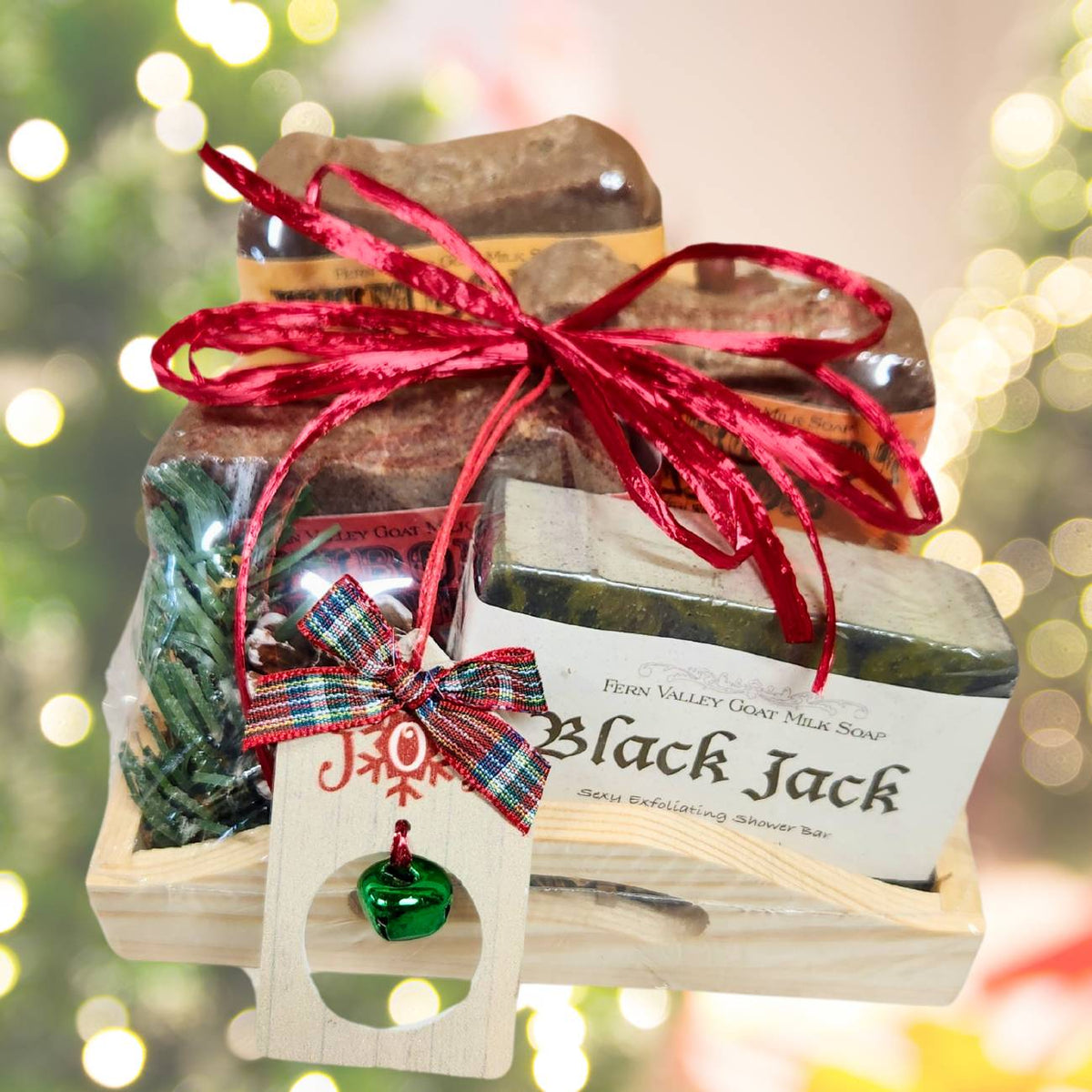 An open wooden gift tray from Fern Valley Soap tied with a festive red ribbon, featuring three stacked Humboldt Hands heavy-duty soaps, a Black Jack goat milk shower bar, pine needles, and a wooden jingle bell holiday tag against a glowing bokeh background.