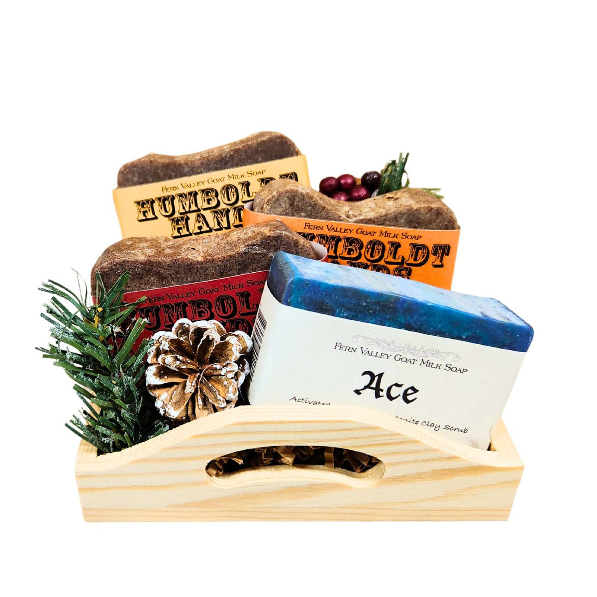 An open wooden gift tray from Fern Valley Soap filled with brown crinkle paper, featuring three stacked Humboldt Hands heavy-duty hand cleaners behind a Ace goat milk shower bar accented with faux pine needles, a frosted pinecone, and festive red berries.