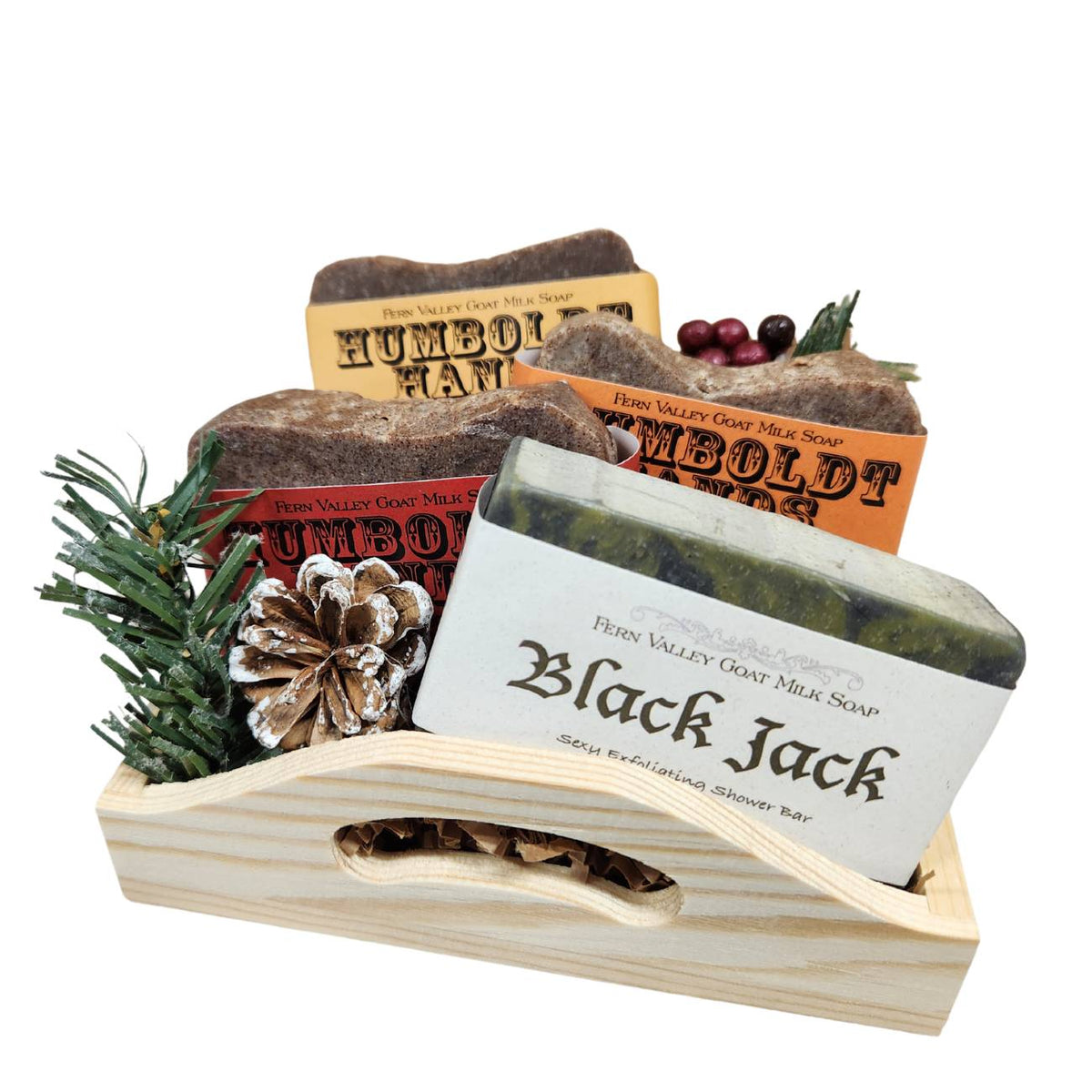 An open wooden gift tray from Fern Valley Soap filled with brown crinkle paper, featuring three stacked Humboldt Hands heavy-duty hand cleaners behind a Black Jack goat milk shower bar accented with faux pine needles, a frosted pinecone, and festive red berries.