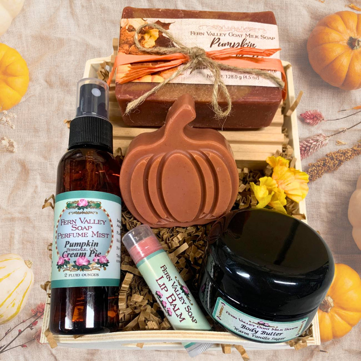 An open wooden gift tray from Fern Valley Soap filled with brown crinkle paper, featuring Pumpkin Apple Butter goat milk soap tied with raffia, a pumpkin novelty soap, Pumpkin Cream Pie perfume mist, a jar of vanilla body butter, and a lip balm, surrounded by mini pumpkins.