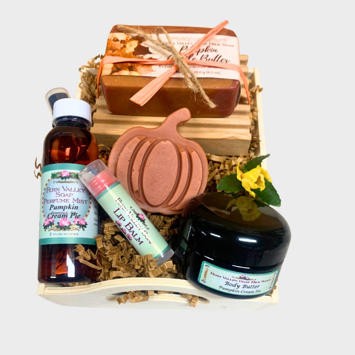An open wooden gift tray from Fern Valley Soap filled with brown crinkle paper, featuring Pumpkin Apple Butter goat milk soap tied with raffia, a pumpkin novelty soap, Pumpkin Cream Pie perfume mist, a jar of vanilla body butter, and a lip balm, on a white background.