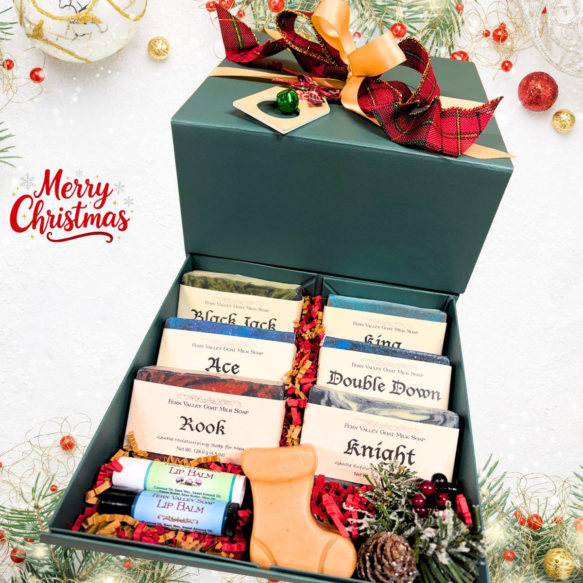 An open pine-green holiday gift box filled with six labeled masculine goat milk soaps, two lip balms, a festive boot-shaped novelty soap, and red shred filler next to Christmas ornaments.