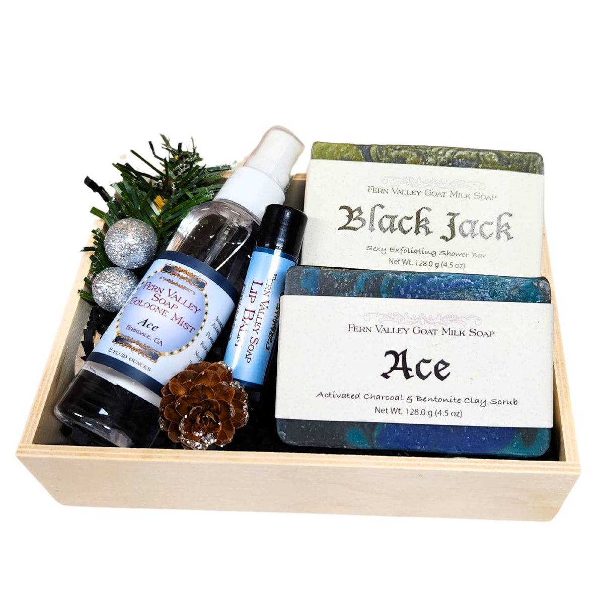 An open wood gift crate for men showcasing two bars of handmade goat milk soap, Black Jack and Ace, alongside a 2-ounce bottle of Ace cologne mist, a tube of lip balm, decorative silver holiday ornaments, and a pinecone.