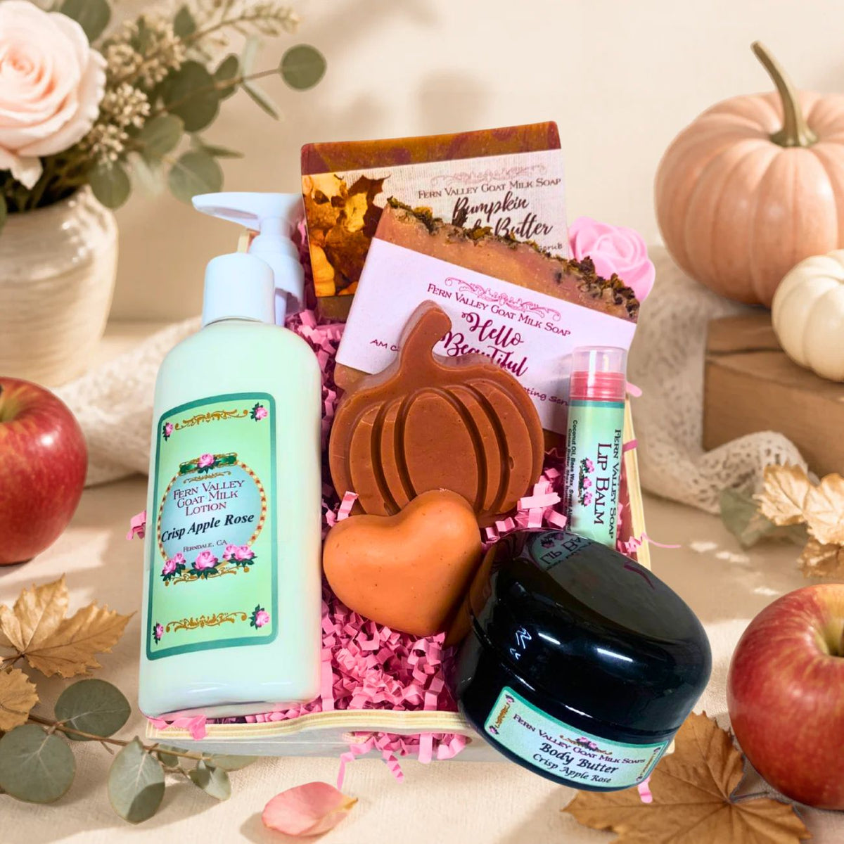 Open autumn gift crate filled with pink crinkle paper, displaying Crisp Apple Rose goat milk lotion, body butter, a pumpkin shaped soap, a mini heart soap, full-size bars, and a lip balm.