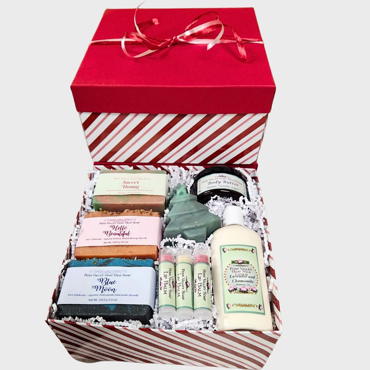 A candy-striped holiday gift box tied with a red bow, filled with Fern Valley Soap products, including three artisan goat milk soaps, body butter, a bottle of lotion, three lip balms, and a Christmas tree-shaped soap.