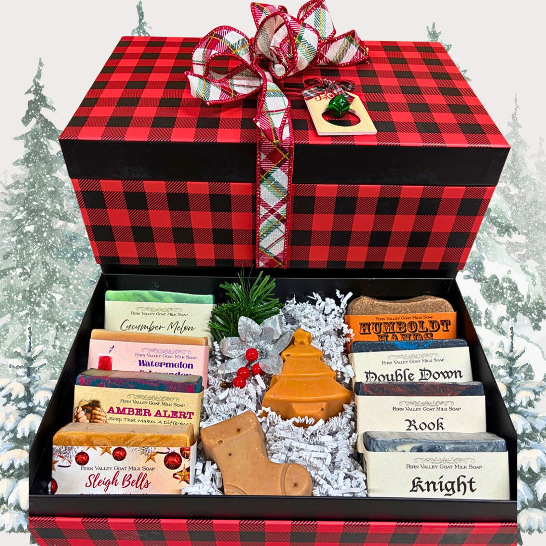 A Christmas-themed gift box filled with assorted soap bars, novelty Christmas-shaped soaps, and small greenery accents. The box is decorated with a red-and-black buffalo plaid pattern and a matching ribbon bow with a small holiday tag.