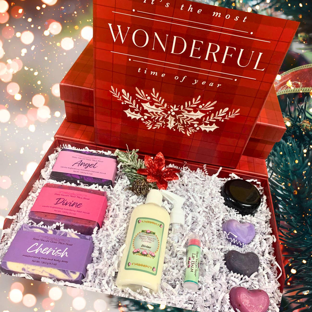 An open red plaid Christmas gift box from Fern Valley Soap reading "Wonderful time of year," filled with white crinkle paper, Angel, Divine, and Cherish goat milk soap bars, a bottle of Divine lotion, a jar of vanilla body butter, a tube of lip balm, and three purple and pink heart soaps.