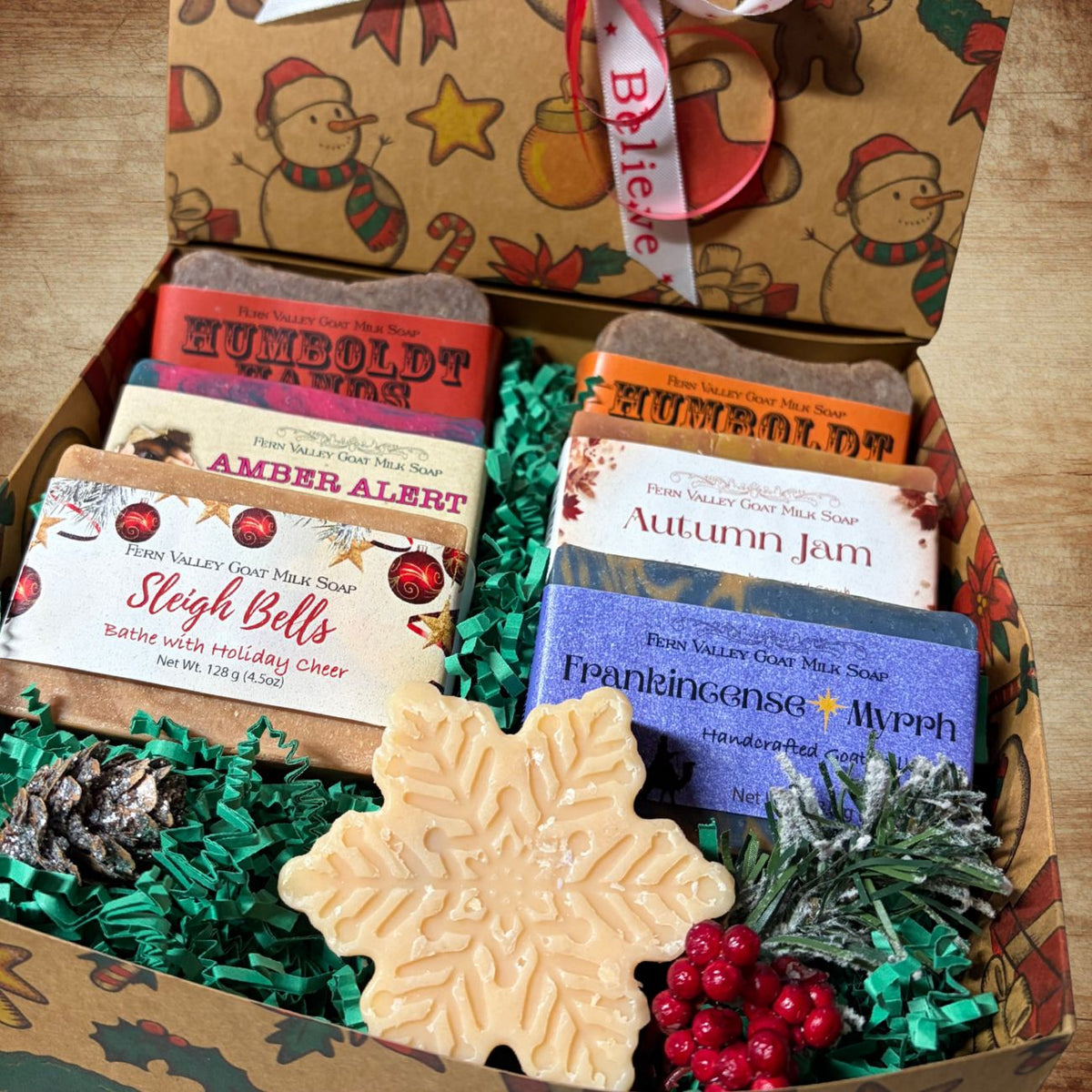 An open gift box filled with handmade soap bars, decorative green shredded paper, pinecones, berries, and a snowflake-shaped soap in the foreground. The box is decorated with a holiday pattern, and the soaps have labels including &