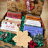 An open gift box filled with handmade soap bars, decorative green shredded paper, pinecones, berries, and a snowflake-shaped soap in the foreground. The box is decorated with a holiday pattern, and the soaps have labels including &