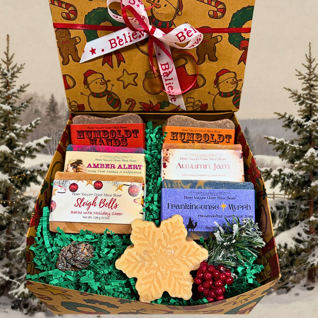 A Christmas-themed gift box filled with assorted soaps, set against a snowy outdoor background with pine trees. The open box contains multiple soap bars in colorful packaging, green shredded paper filler, a large snowflake-shaped soap, red decorative berries, and pinecones. 