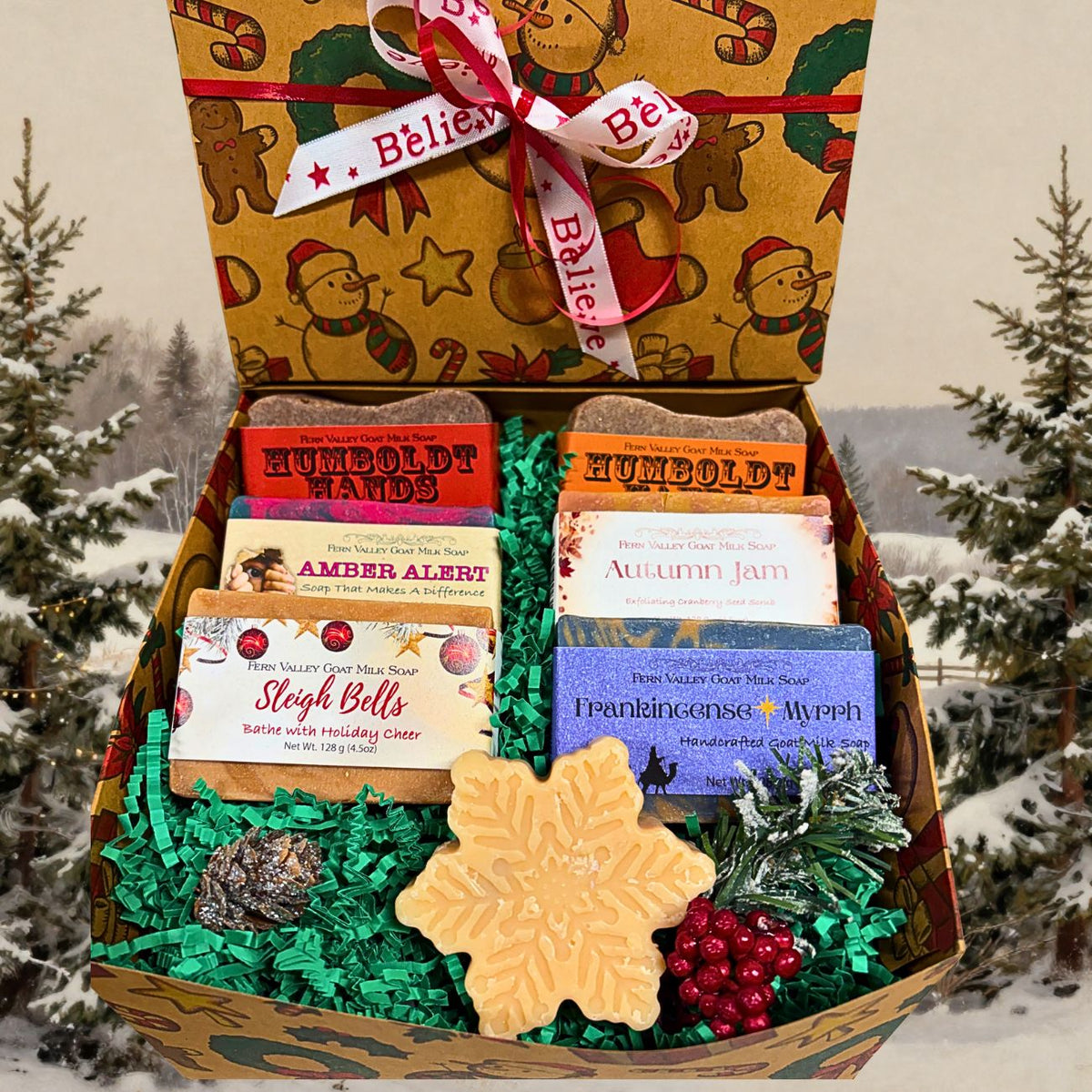 A Christmas-themed gift box filled with assorted soaps, set against a snowy outdoor background with pine trees. The open box contains multiple soap bars in colorful packaging, green shredded paper filler, a large snowflake-shaped soap, red decorative berries, and pinecones. 