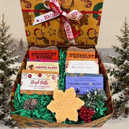 A Christmas-themed gift box filled with assorted soaps, set against a snowy outdoor background with pine trees. The open box contains multiple soap bars in colorful packaging, green shredded paper filler, a large snowflake-shaped soap, red decorative berries, and pinecones. 
