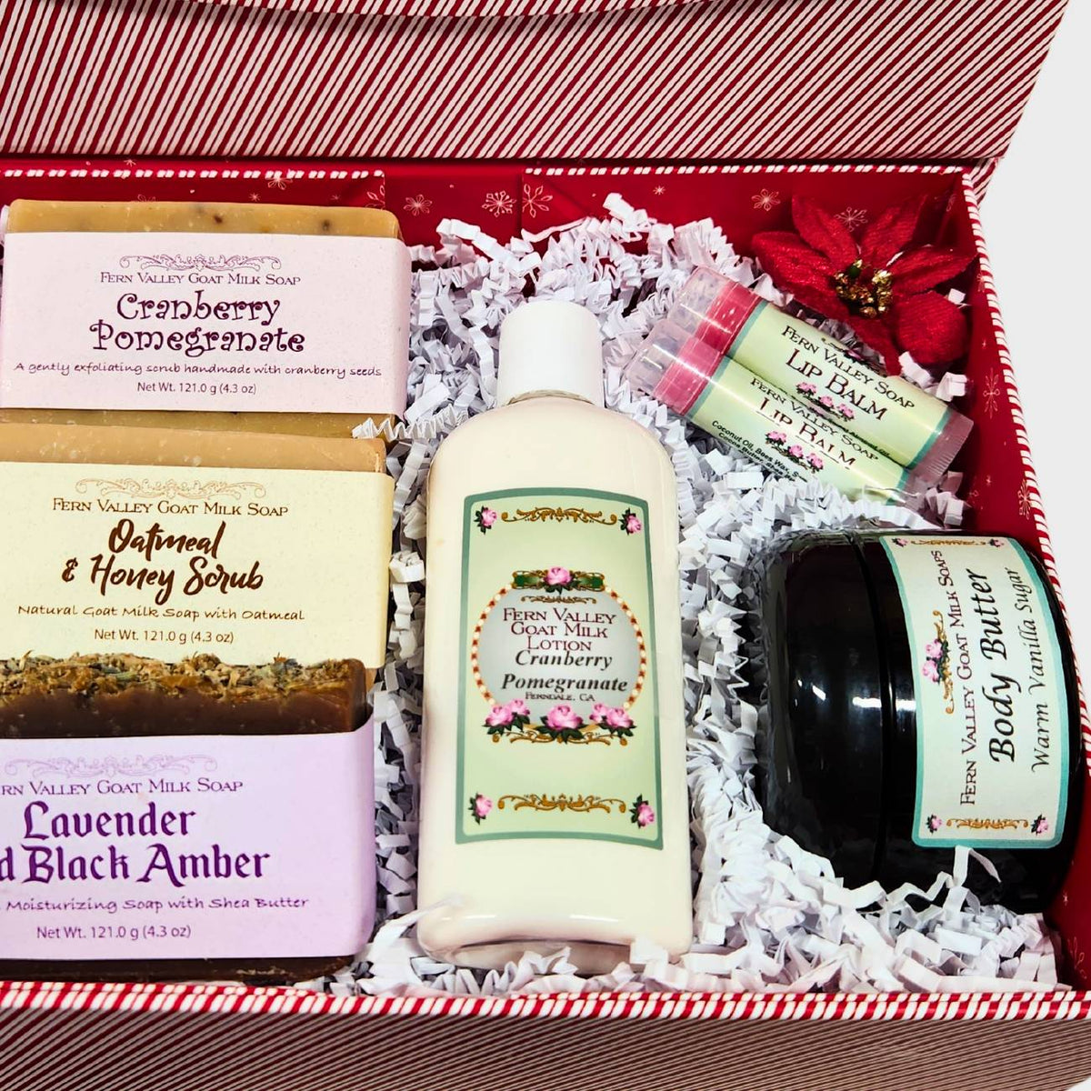 An open red and white striped Christmas gift box , neatly filled with white crinkle paper, three Fern Valley goat milk soap bars (Cranberry Pomegranate, Oatmeal &amp; Honey, Lavender and Black Amber), a bottle of lotion, a jar of body butter, and two lip balms.