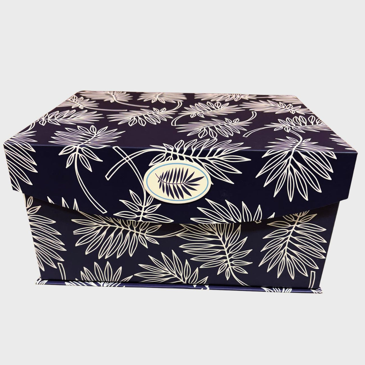 A rectangular gift box with a navy blue base and a white tropical palm leaf pattern, featuring a circular gold-toned logo medallion on the lid, set against a plain light gray background.
