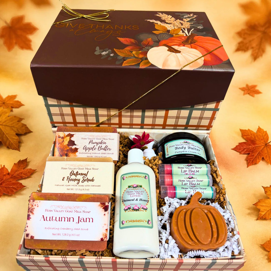 Open Give Thanks Always plaid gift box filled with white crinkle paper, displaying pumpkin apple butter, oatmeal, and autumn jam goat milk soaps, lotion, body butter, a pumpkin shaped soap, and three lip balms.