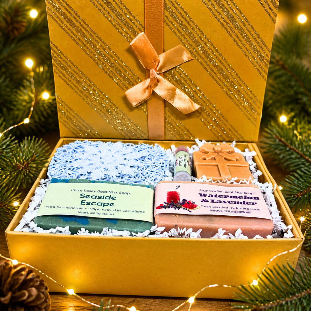 Open gold glitter holiday gift box with Seaside Escape and Watermelon &amp; Lavender goat milk soaps, a lip balm, and a present-shaped soap novelty, nestled in blue paper shred with warm fairy lights and evergreen pine branches in the background.