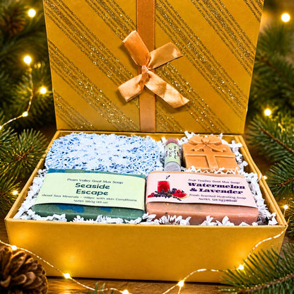 Open gold glitter holiday gift box with Seaside Escape and Watermelon &amp; Lavender goat milk soaps, a lip balm, and a present-shaped soap novelty, nestled in blue paper shred with warm fairy lights and evergreen pine branches in the background.
