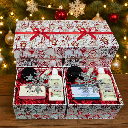 Two open customizable holiday gift boxes displaying the Queen and blue artisan soap gift sets with matching lotions and body jars, sitting below a stacked matching closed box with a red ribbon against a glowing, festive Christmas tree bokeh background.