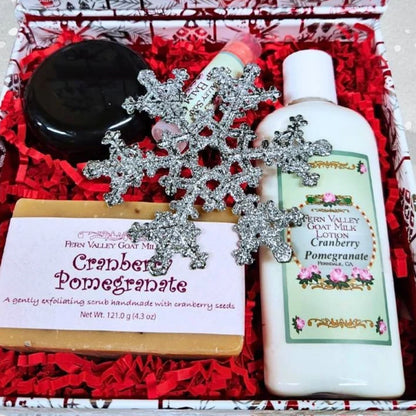 A gift set in a red patterned box filled with red shredded paper, containing a bar of Fern Valley Cranberry Pomegranate soap, a bottle of Fern Valley Goat Milk Lotion Cranberry Pomegranate, a jar of body butter, and a lip balm. Includes a silver snowflake ornament.