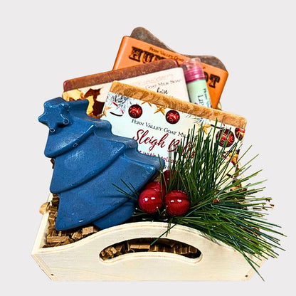 An open wooden gift tray filled with handmade goat milk soaps, including Humboldt Hands, Pumpkin Apple Butter, and Sleigh Bells. A deep blue, tree-shaped novelty soap stands in the foreground next to decorative pine needles, winter berries, and a pink tube of lip balm.