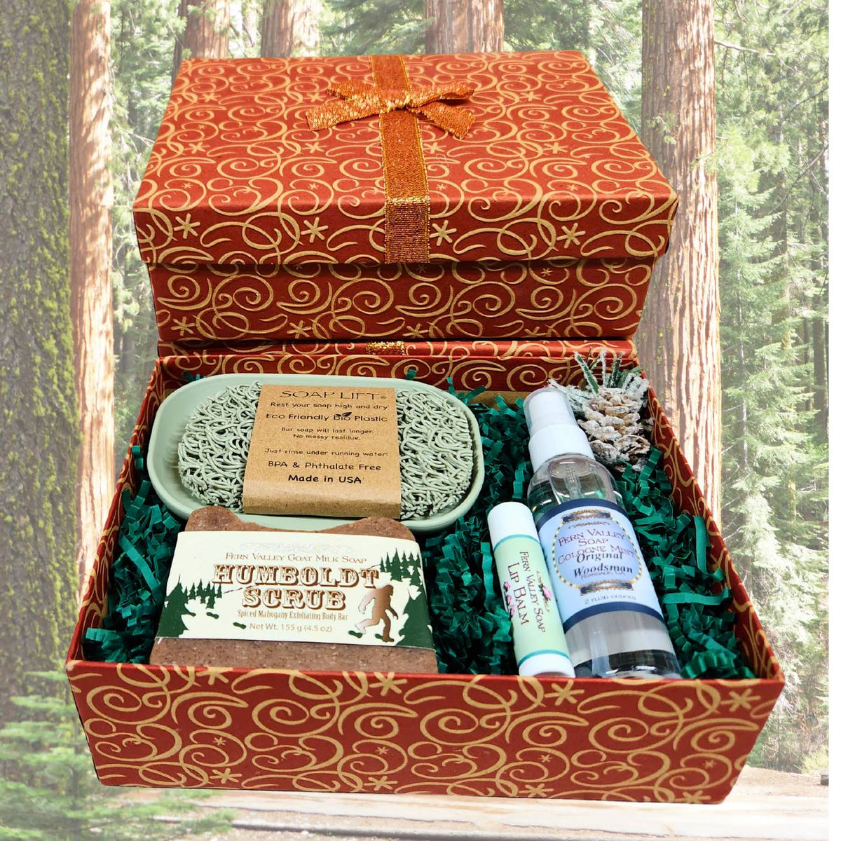 An open copper and gold patterned Christmas gift box filled with green crinkle paper, displaying a Humboldt Scrub goat milk soap bar, a 2 oz Original Woodsman cologne spray, a lip balm, and a sage green self-draining soap dish with a matching soap lift. The background is redwood trees.