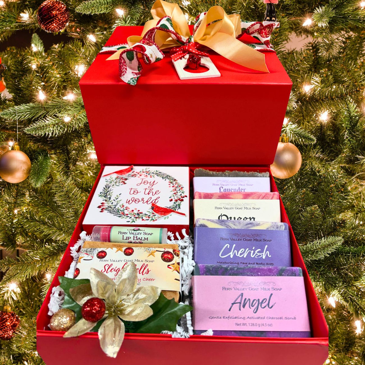 An open luxury red Christmas gift box filled with five labeled Fern Valley goat milk soaps (Sleigh Bells, Lavender, Queen, Cherish, Angel), a lip balm, and a winter bird wooden plaque, styled in front of a  lit Christmas tree.