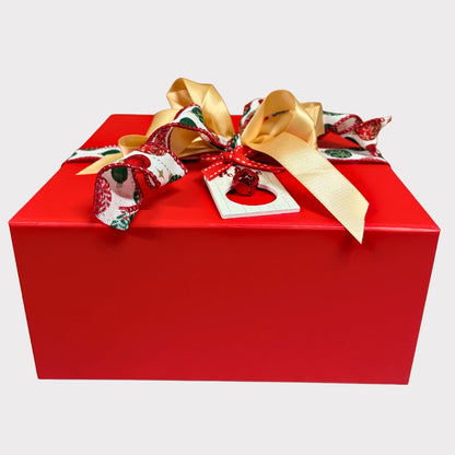 A closed square crimson red gift box elegantly tied with a gold satin bow, a red and green holly patterned ribbon, and a tiny red jingle bell ornament tag against a white background.