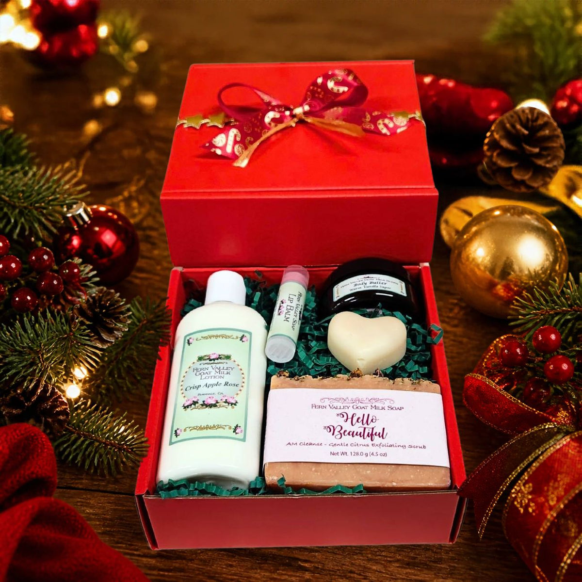 An open holiday red gift box from Fern Valley Soap tied with a red and gold ribbon, featuring Hello Beautiful goat milk soap, Crisp Apple Rose lotion, a heart novelty soap, vanilla body butter, and a lip balm on green crinkle paper.