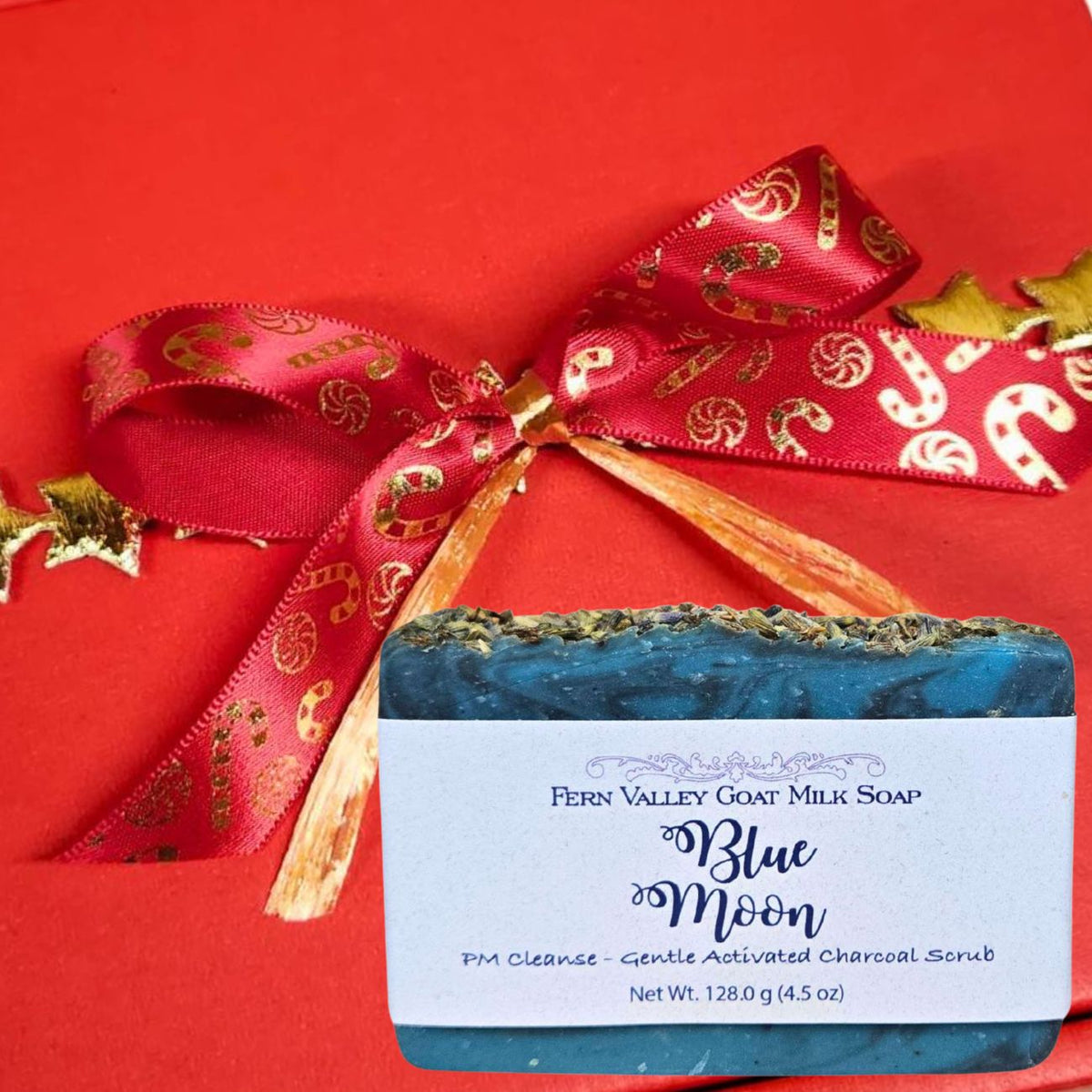 A bar of Fern Valley Goat Milk Soap labeled Blue Moon. The background is a solid red box tied with a red ribbon printed with gold candy canes.