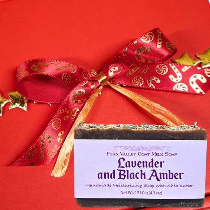 A bar of Fern Valley Goat Milk Soap labeled Lavender Black Amber. The background is a solid red box tied with a red ribbon printed with gold candy canes.