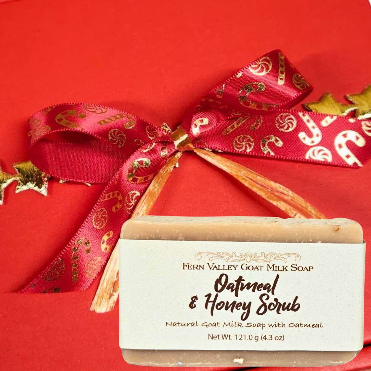 A bar of Fern Valley Goat Milk Soap labeled Oatmeal &amp; Honey. The background is a solid red box tied with a red ribbon printed with gold candy canes.
