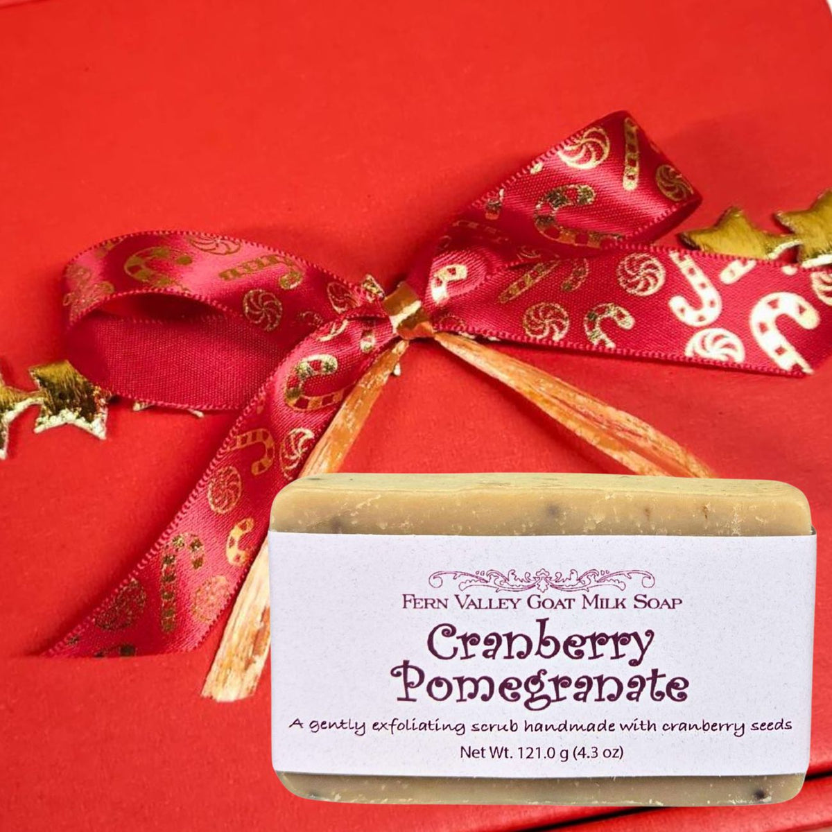 A bar of Fern Valley Goat Milk Soap labeled Cranberry Pomegranate. The background is a solid red box tied with a red ribbon printed with gold candy canes.