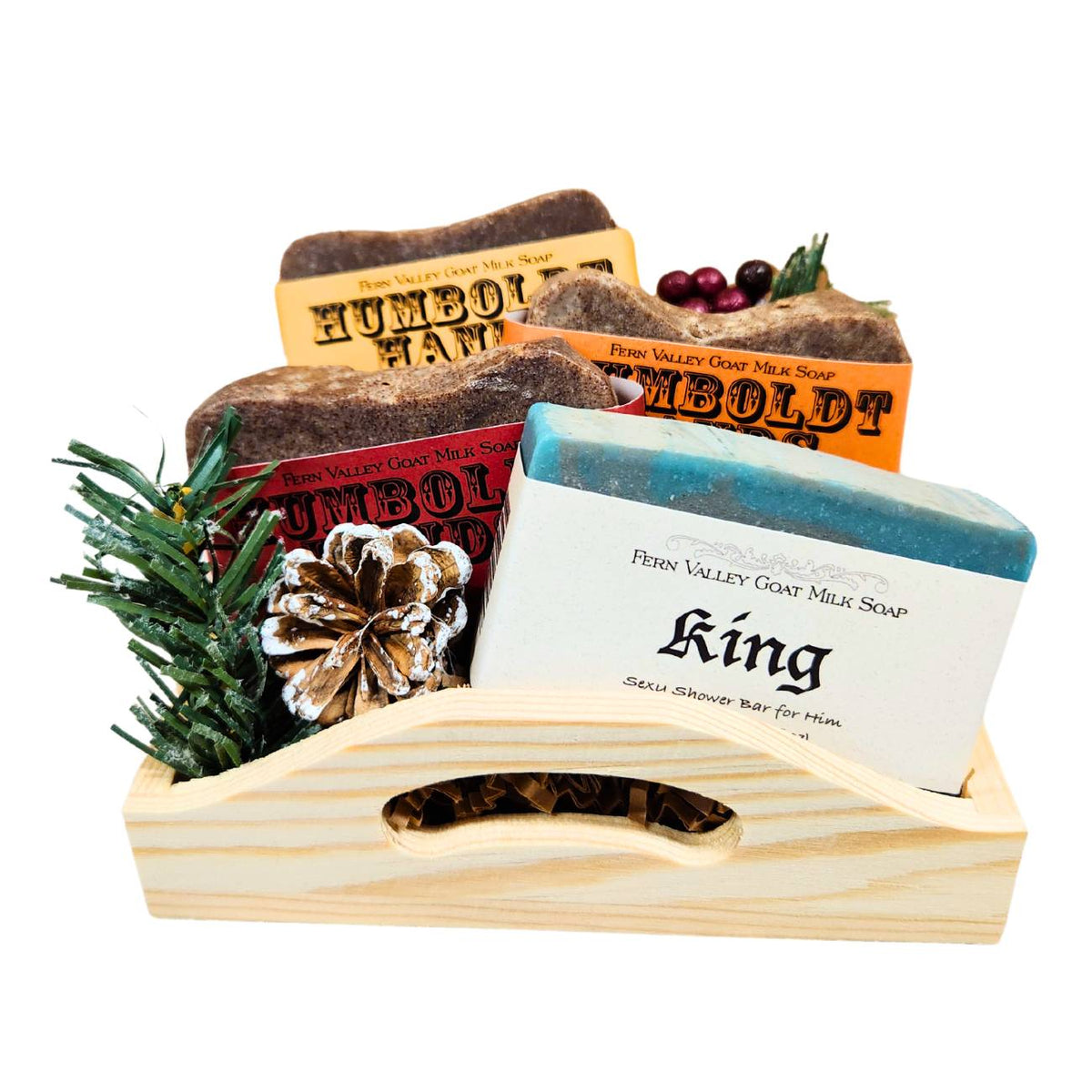 An open wooden gift tray from Fern Valley Soap filled with brown crinkle paper, featuring three stacked Humboldt Hands heavy-duty hand cleaners behind a King goat milk shower bar with a striking blue and gray swirl, accented with faux pine needles, a frosted pinecone, and festive red berries.