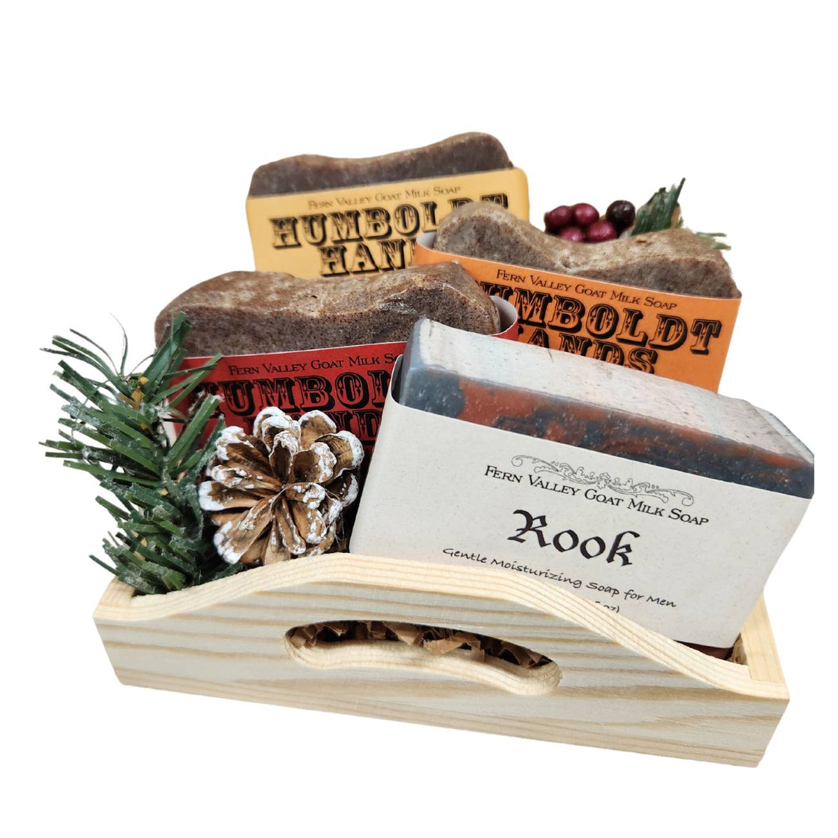 An open wooden gift tray from Fern Valley Soap filled with brown crinkle paper, featuring three stacked Humboldt Hands heavy-duty hand cleaners behind a Rook goat milk shower bar accented with faux pine needles, a frosted pinecone, and festive red berries.