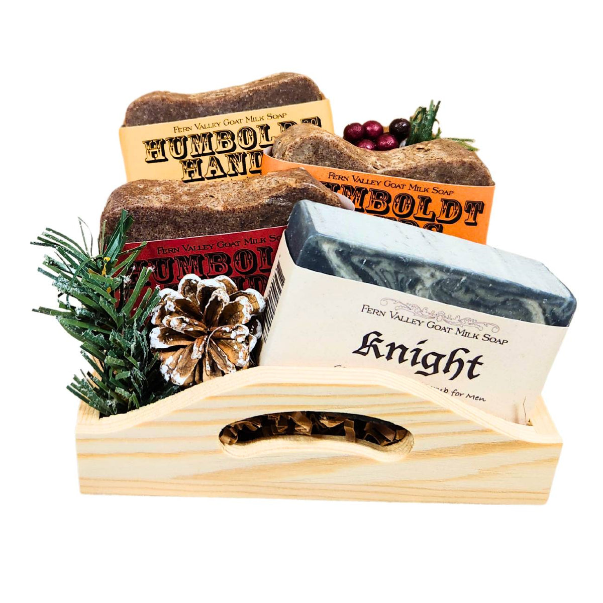 An open wooden gift tray from Fern Valley Soap filled with brown crinkle paper, featuring three stacked Humboldt Hands heavy-duty hand cleaners behind a Knight goat milk shower bar accented with faux pine needles, a frosted pinecone, and festive red berries.