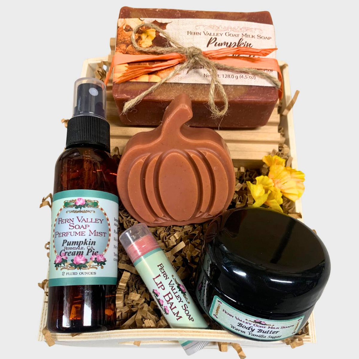 An open wooden gift tray from Fern Valley Soap filled with brown crinkle paper, featuring Pumpkin Apple Butter goat milk soap tied with raffia, a pumpkin novelty soap, Pumpkin Cream Pie perfume mist, a jar of vanilla body butter, and a lip balm, on a white background.