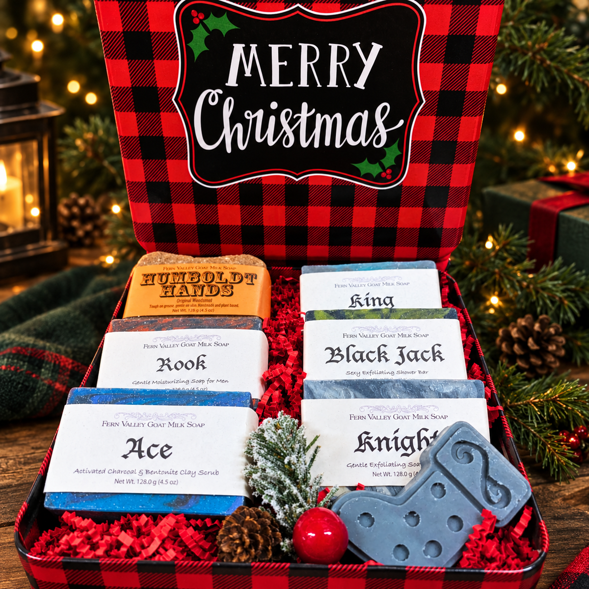 A Christmas-themed gift set in a red and black buffalo plaid metal box, featuring six different colored soap bars, a gray stocking-shaped soap, a small pine sprig, a red ornament, and pinecones, with a red and black plaid lid printed with &