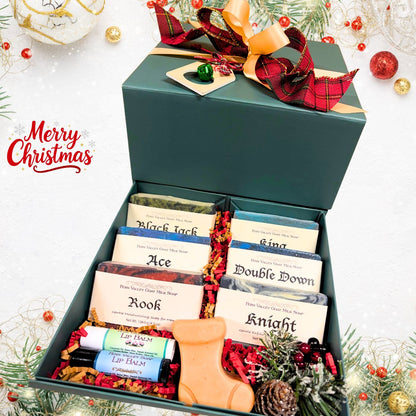An open pine-green holiday gift box filled with six labeled masculine goat milk soaps, two lip balms, a festive boot-shaped novelty soap, and red shred filler next to Christmas ornaments.