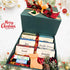 An open pine-green holiday gift box filled with six labeled masculine goat milk soaps, two lip balms, a festive boot-shaped novelty soap, and red shred filler next to Christmas ornaments.