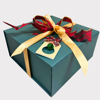 A closed luxury pine-green magnetic gift box elegantly tied with a gold satin bow, a red plaid ribbon, and a miniature green jingle bell charm tag against a clean white background.