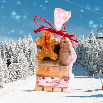 A packaged holiday gift set on a snowy mountain path, featuring gingerbread-themed soap shaped like a gingerbread man and a snowman, wrapped in clear cellophane with a red ribbon. The front soap label reads &